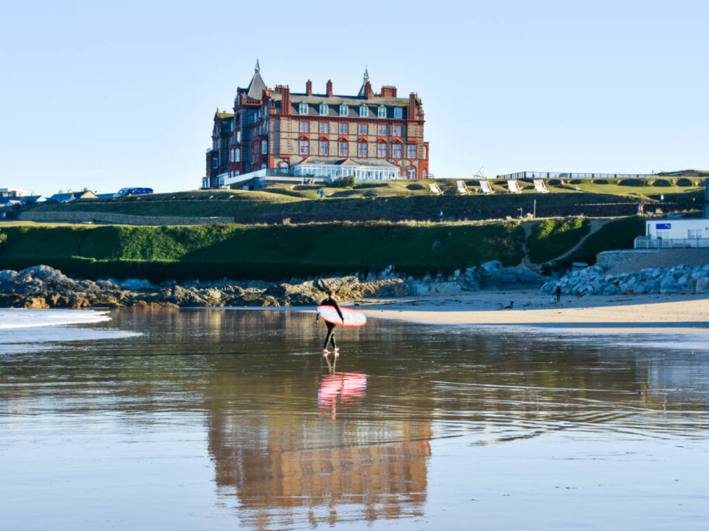 A surfer walking at the beach with the Headland Hotel & Spa in the background