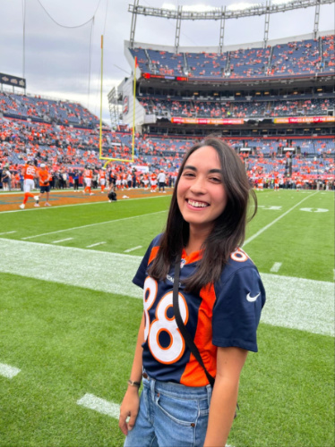 The author Abigail posing for a photo at the field with the Broncos in the background in Empower Field at Mile High