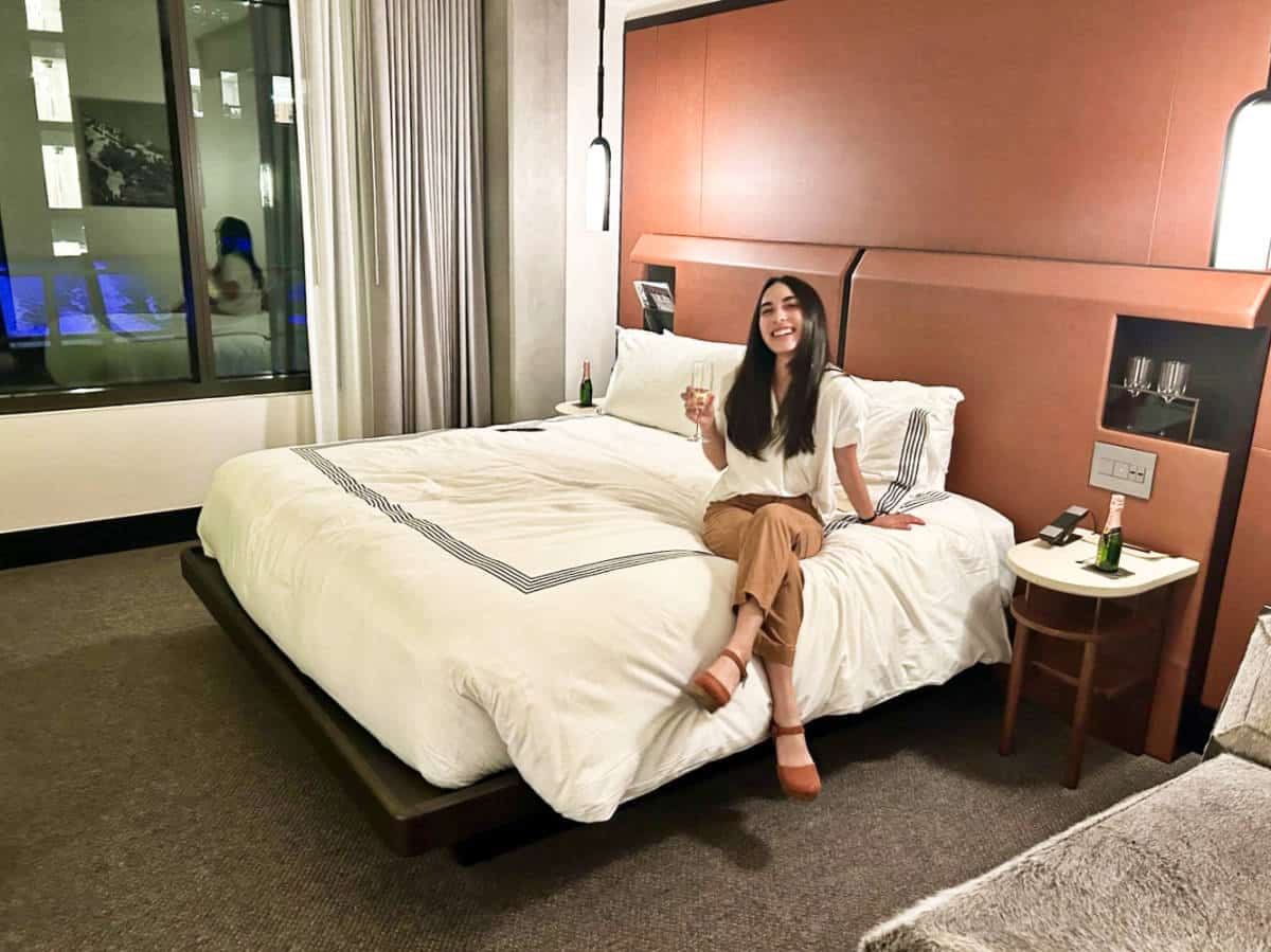 Travel Lemming editor Abigail Bliss holding a glass of champagne while sitting on the bed in Thompson Denver Hotel