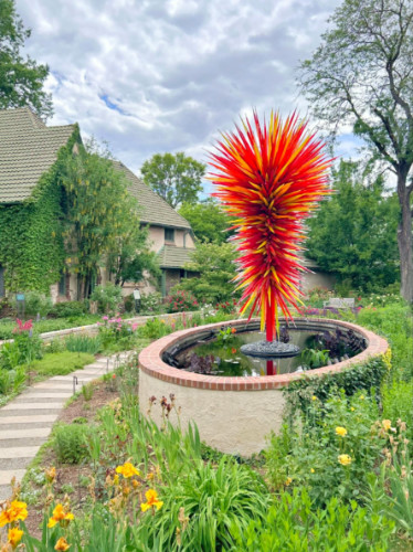 A red and yellow art sculpture resembling a spiky burst stands at the center of a circular pond at the Denver Botanic Gardens