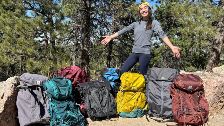 The author Abigail Bliss showing the 8 backpacks that she tested and compared, in a wooded trail