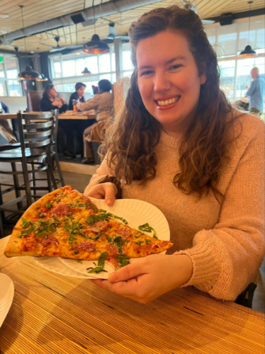 Author Elizabeth Demolat showing a slice of pizza on a paper plate at Five Points Pizza