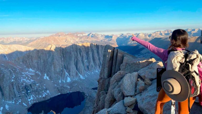 Author McKenna stands atop a mountain, wearing a backpack, enjoying the breathtaking view from the summit.