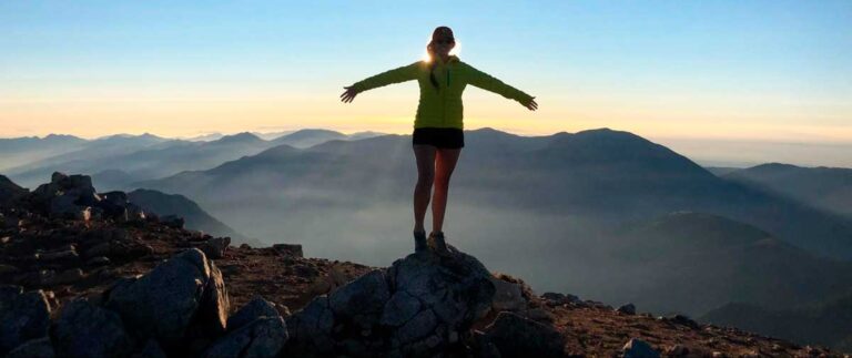 Author McKenna stands on a mountain summit, arms wide open, enjoying the panoramic view