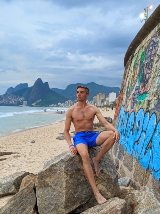 Author Nate in blue trunks sits on a rock by the beach, gazing at the ocean waves