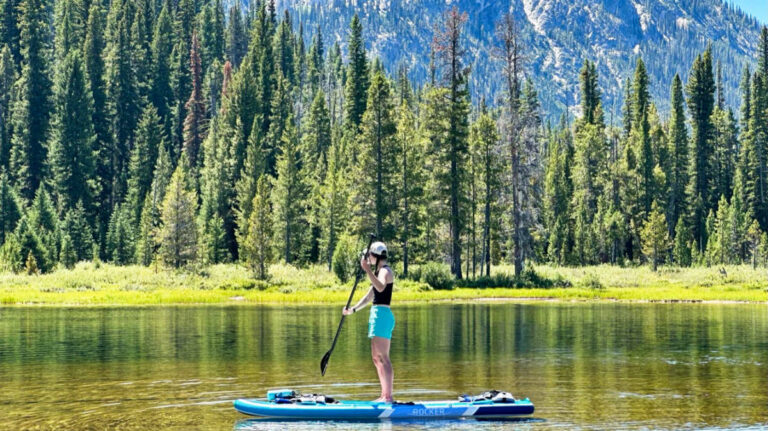 The author Taylor Ramsey, paddleboarding with a picturesque view in the background on a lake