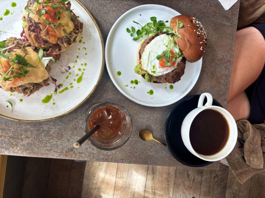 A plate with grilled halloumi, poached eggs & avo on toast and other plate with eggs benedict and drinks at The Farmer’s Mistress