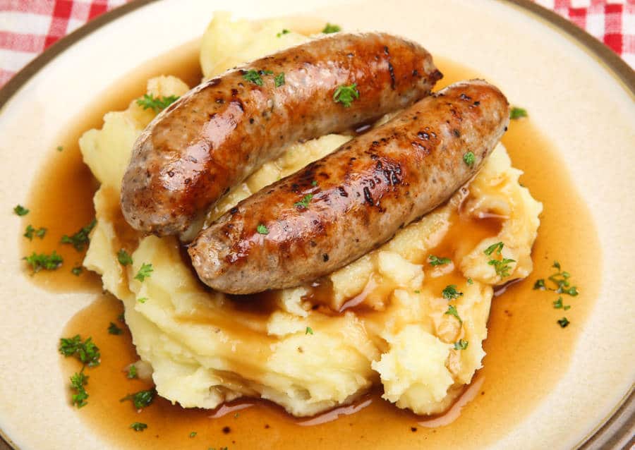 A plate of mashed potato topped with two sausages and gravy