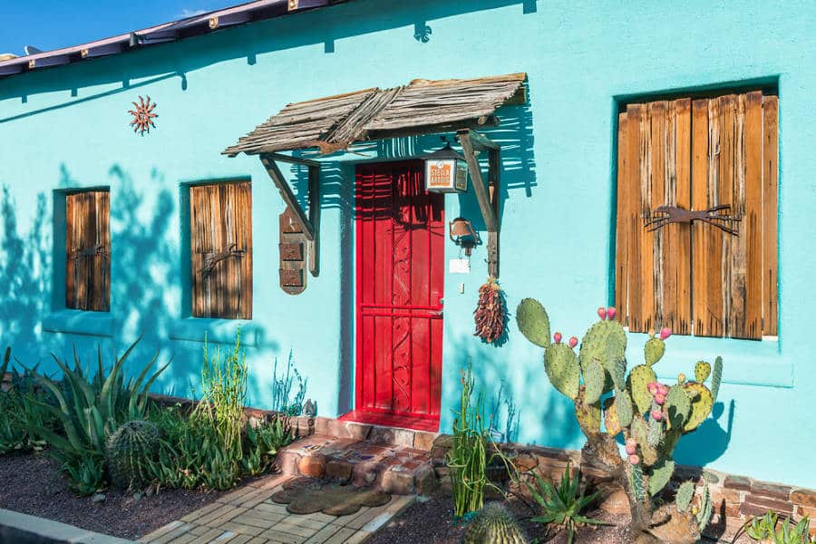 A vibrant turquoise exterior and red door of a home in Barrio Viejo with cacti on the ground
