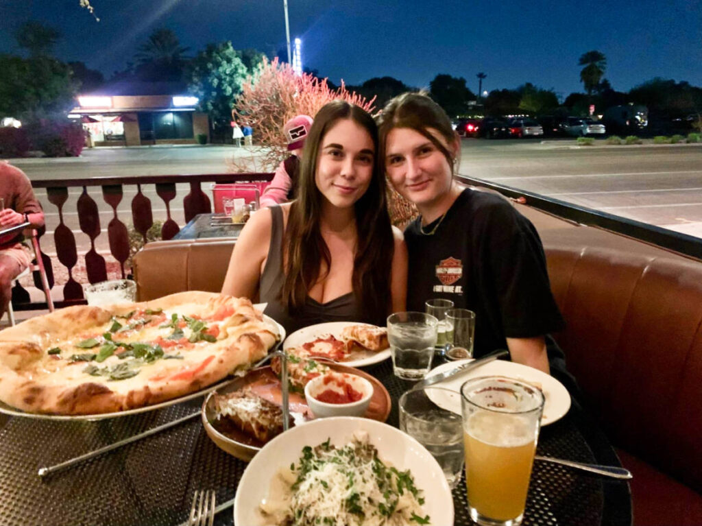 The author Betty Hurd and her best friend, posing for a photo with their served pizza, pasta, and beer at the outdoor patio of Federal Pizza