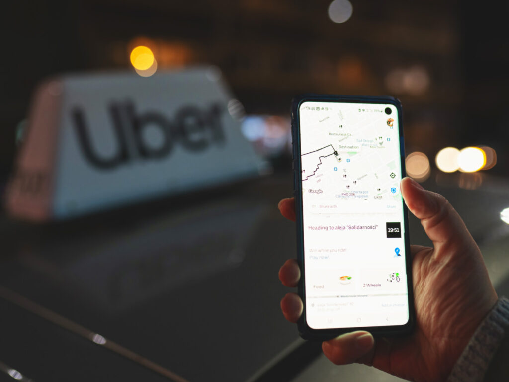 Uber app displayed on smartphone held in hand in front of Uber taxi sign
