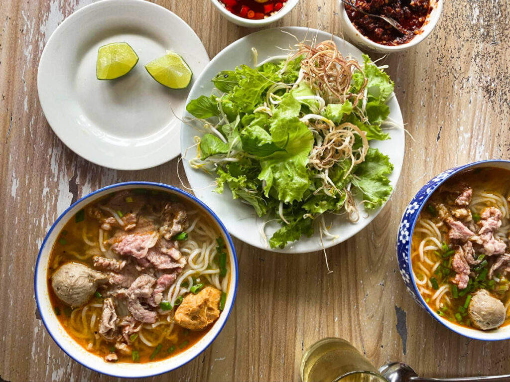 Two bowls of Bún bò Huế, a beef and pork noodle soup with leafy greens on the side