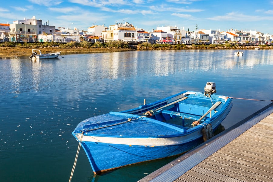 A vibrant blue boat docked on the side of a lake with a cityscape view of Cabanas de Tavira in the background
