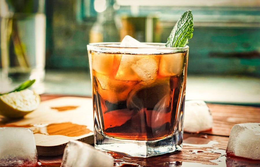 A glass of rum full of ice with a mint leaf on a wooden board with melted ice