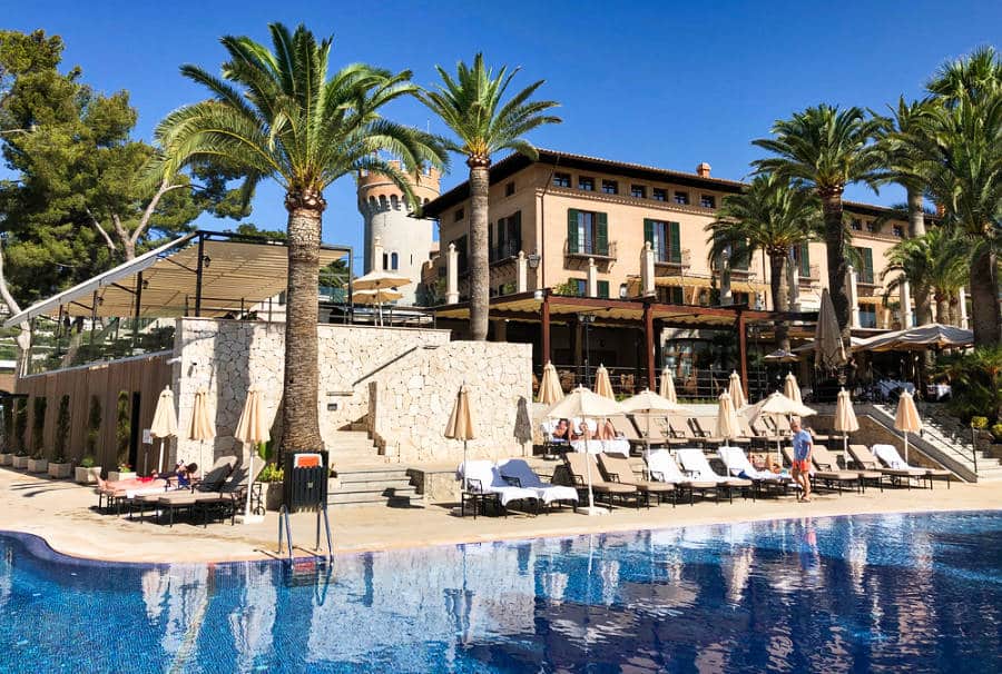 A sunny exterior view of Castillo Hotel Son Vida from the pool surrounded by lounge chairs and palm trees