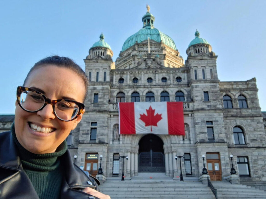 The author Catherine Driver smiling for a photo in front of the BC’s Parliament Buildings with a flag of Canada