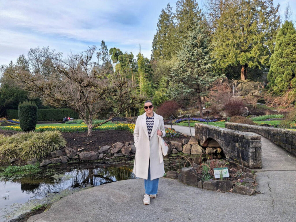 The author Catherine posing for a photo near a small lake surrounded by greenery at Queen Elizabeth Park