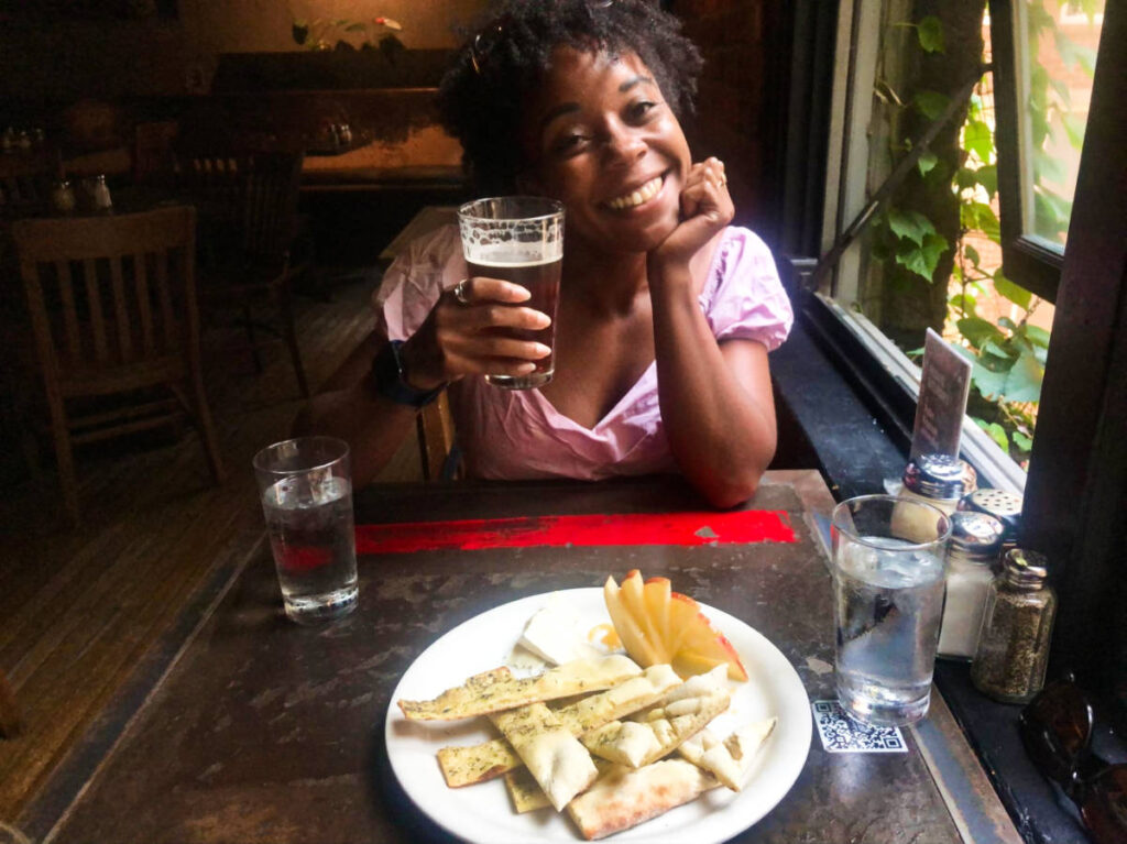The author Chelsea Booker, smiling for a photo while holding a glass of beer with a plate of brie, focaccia & apples on the table