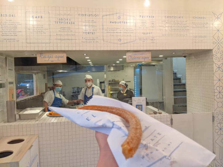A hand holding a churro in front of Churrería El Moro