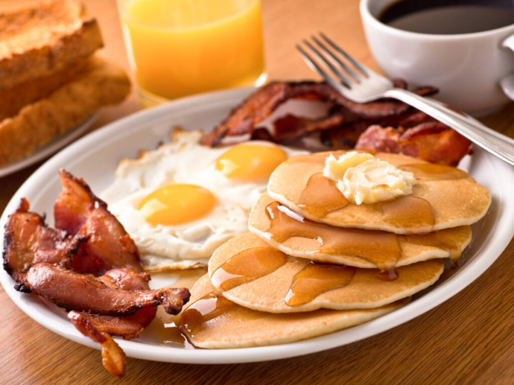 A plate of crispy bacon, eggs, pancakes, toast, coffee, with a glass of orange juice and coffee