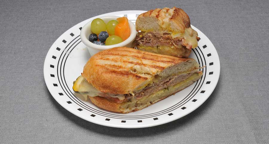 Cuban sandwiches are sliced in halves with a small bowl of different fruits on a plate.