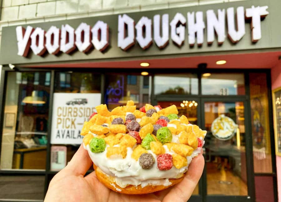 A hand holding a donut topped with colorful cereal in front of Voodoo Doughnut