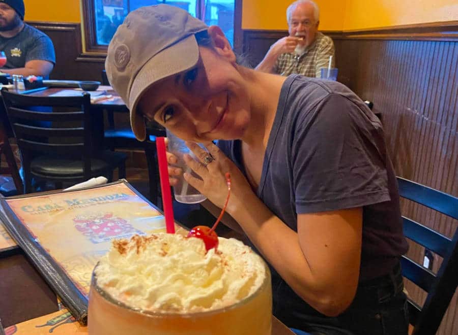 A tall glass of drink with whipped cream and cherry on top in front of a woman at a restaurant.