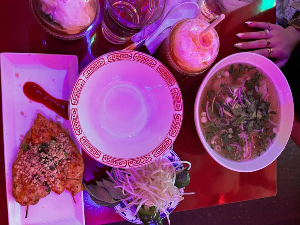 A table full with charboiled chicken skewers, chicken pho, and Thai tea