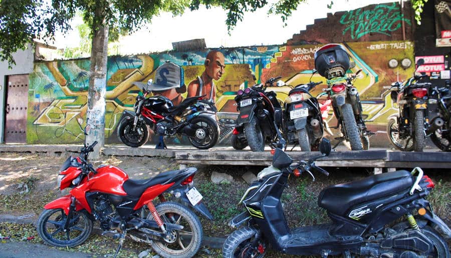 A wall with graffiti and unorganized motorcycles parked at the side of the street