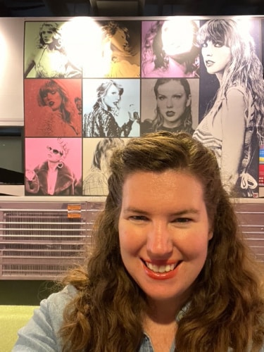 The author, Elizabeth Demolat smiling for a selfie with a poster of Taylor Swift on her background in Country Music Hall of Fame