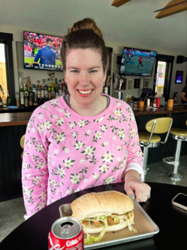 The author Elizabeth Demolat, smiling for a photo with her sandwich and soda at Spicy Boy’s