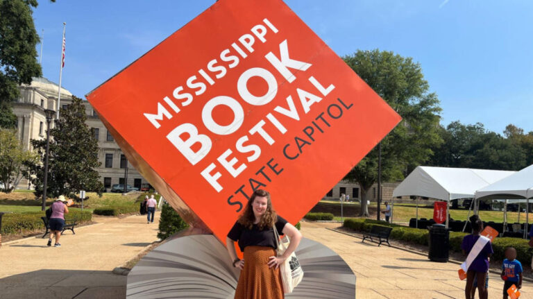 The author Elizabeth Demolat, posing for a photo in front of the landmark signage at the Mississippi Book Festival