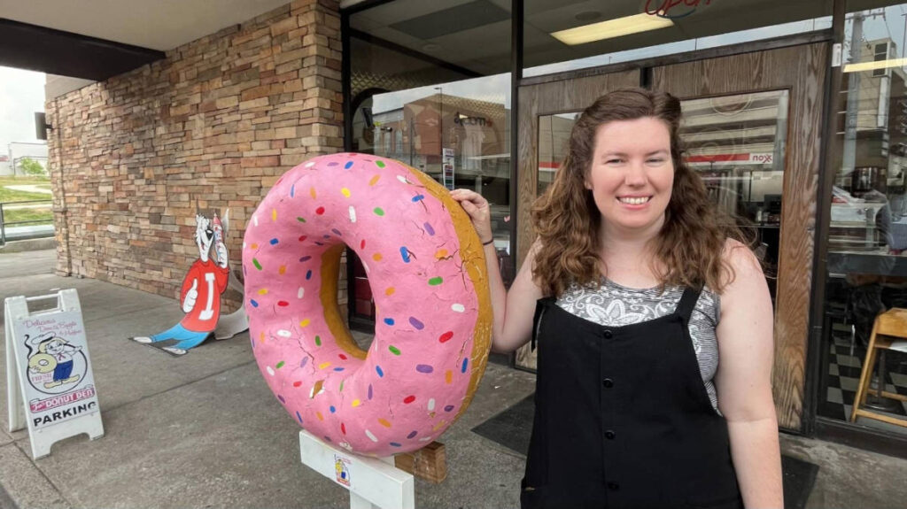 Author Betty posing for a photo with a large donut outside Fox’s Donut Den