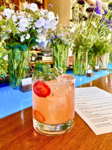 A glass of Fresca Flaca cocktail and garden-variety garnishes in the background