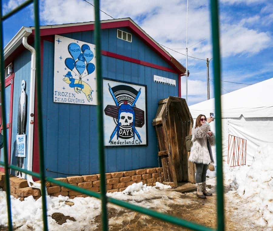 A woman taking a selfie in front of a coffin and blue shed at Frozen Dead Guy Days