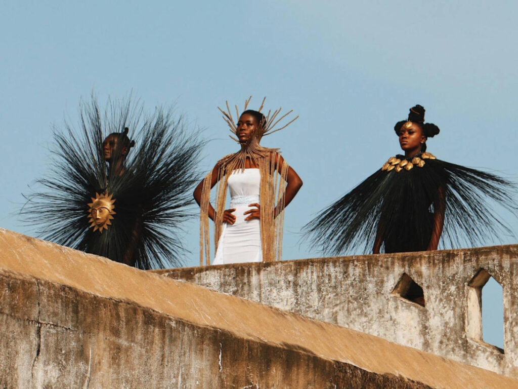 Three Ghanaian models in stylish dresses standing on a wall at Elmina Castle