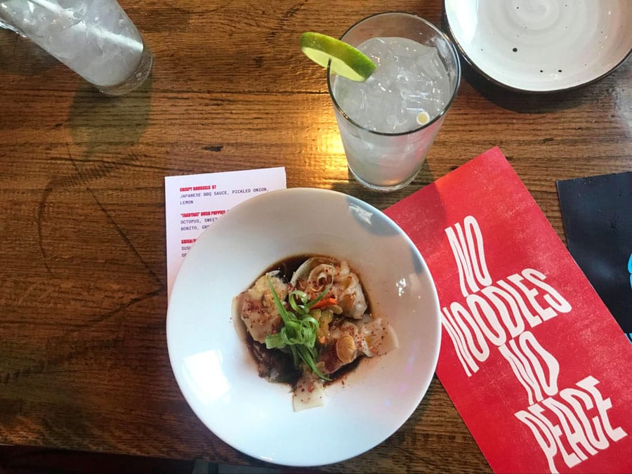 A bowl of dumplings with noodles and a glass of drink with lime in Glo Noodle House