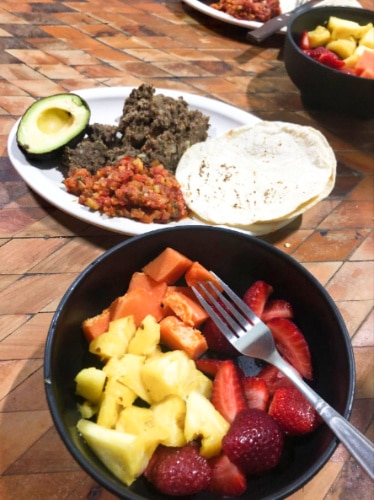 A plate with homemade tacos with a slice of avocado and a bowl of different fruits