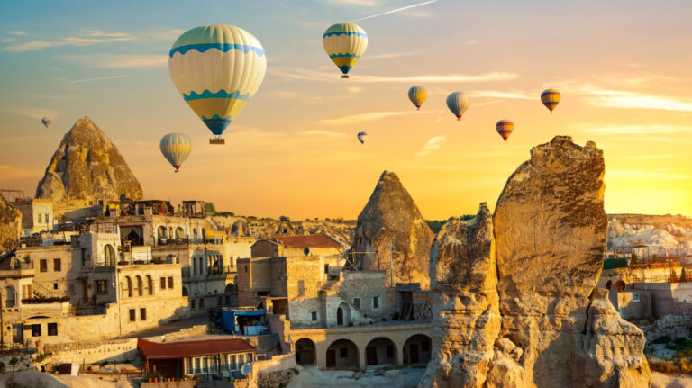 Hot air balloons hover over rock pillars and stone buildings at sunset in Cappadocia
