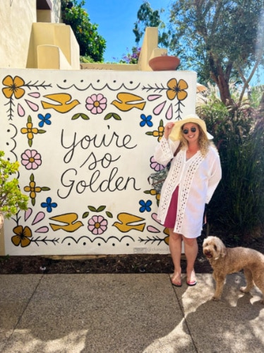 The author Jess Levy and her dog posing outside near the mural of Mission Estancia La Jolla