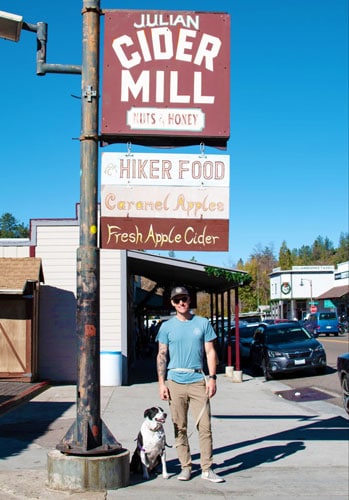 A man with his dog standing beside the Julian Cider Mill signage