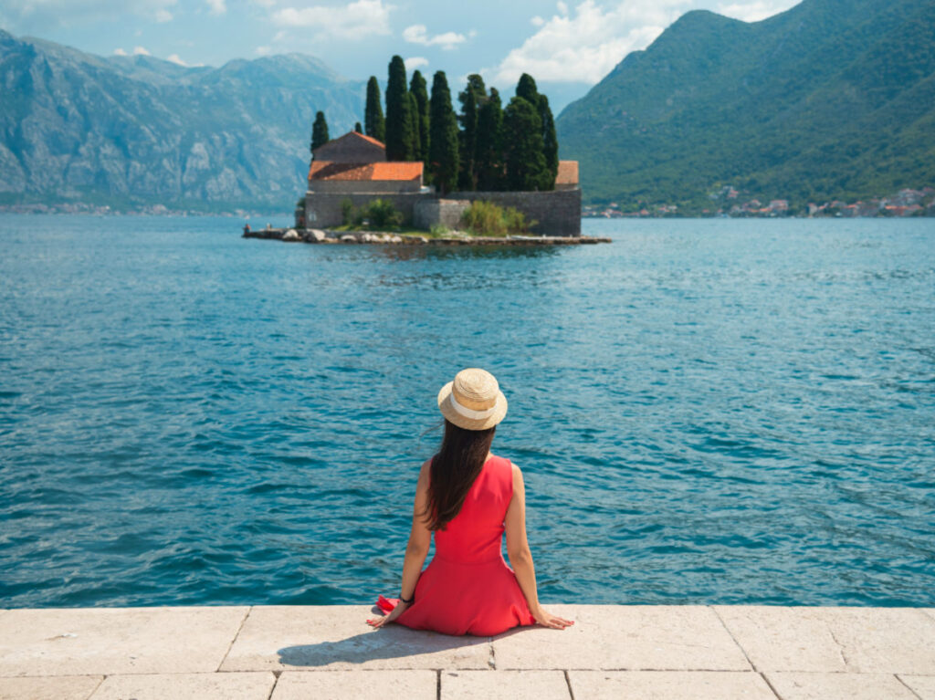 Woman in red dress and hat sitting in front of scenic bay in Perast