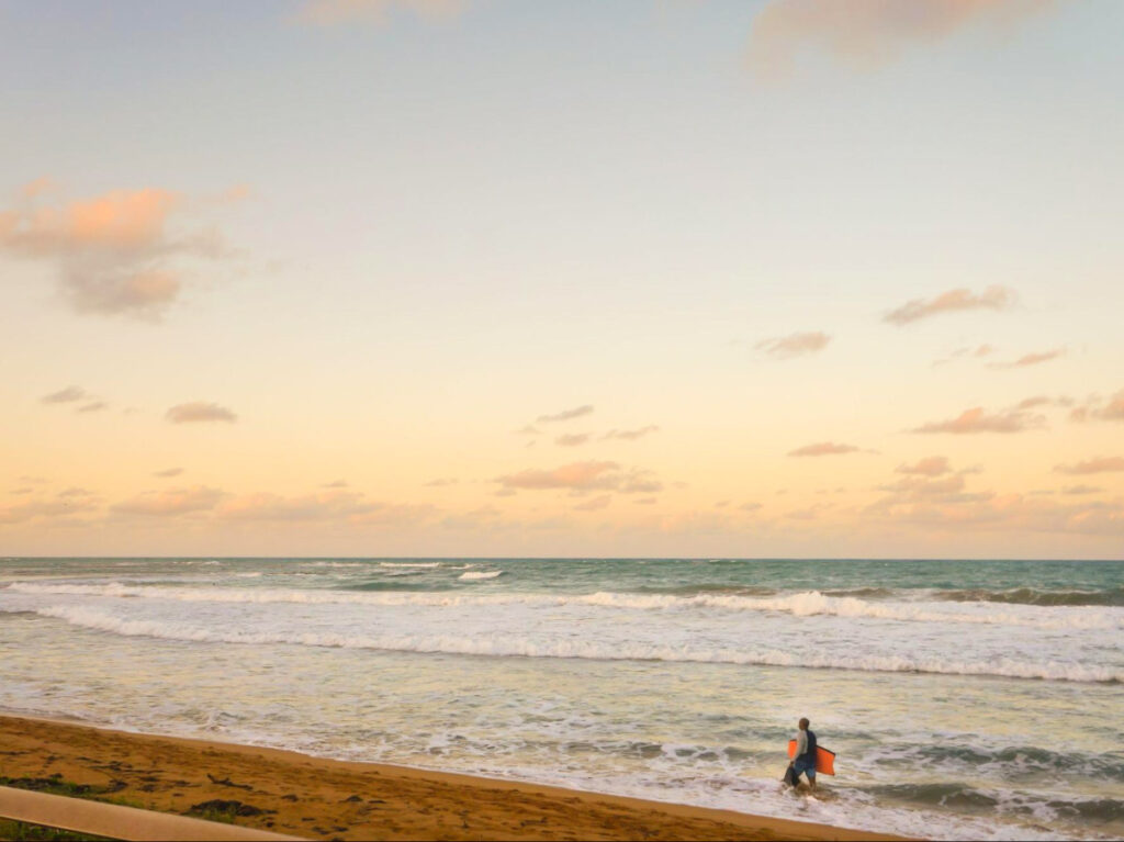 A person walking along La Pared beach in Luquillo with a surfboard