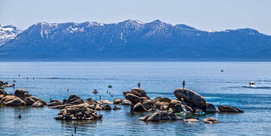 View of people climbing on the rocks, swimming and paddleboarding on the water of Lake Tahoe with a snow tipped mountain view in the distance