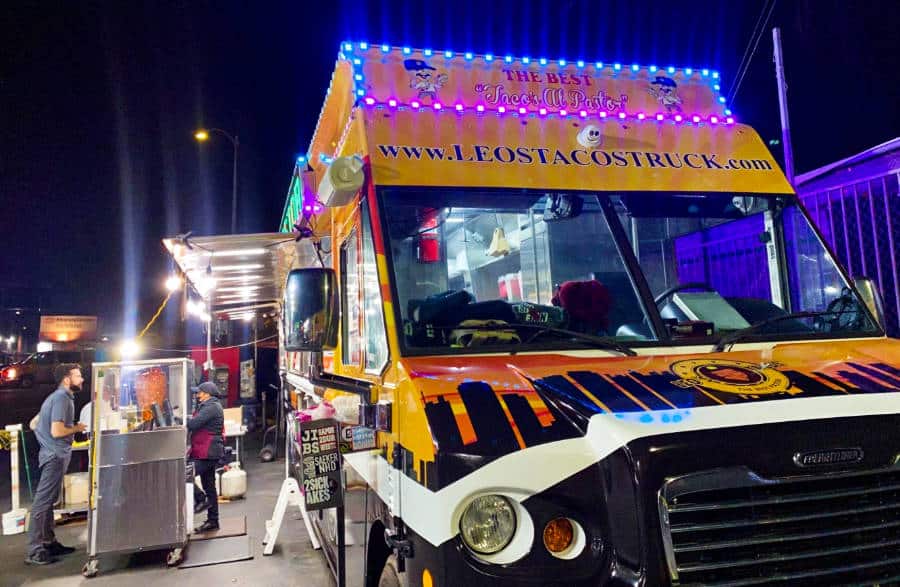 A big yellow truck with neon lights of Leo’s Taco Truck parked ready to serve in the street of Los Angeles