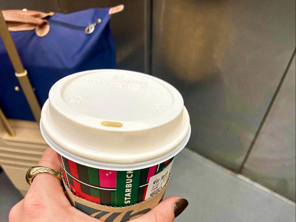 A hand holding a cup of coffee from Starbucks with a suitcase, and day bag in the background