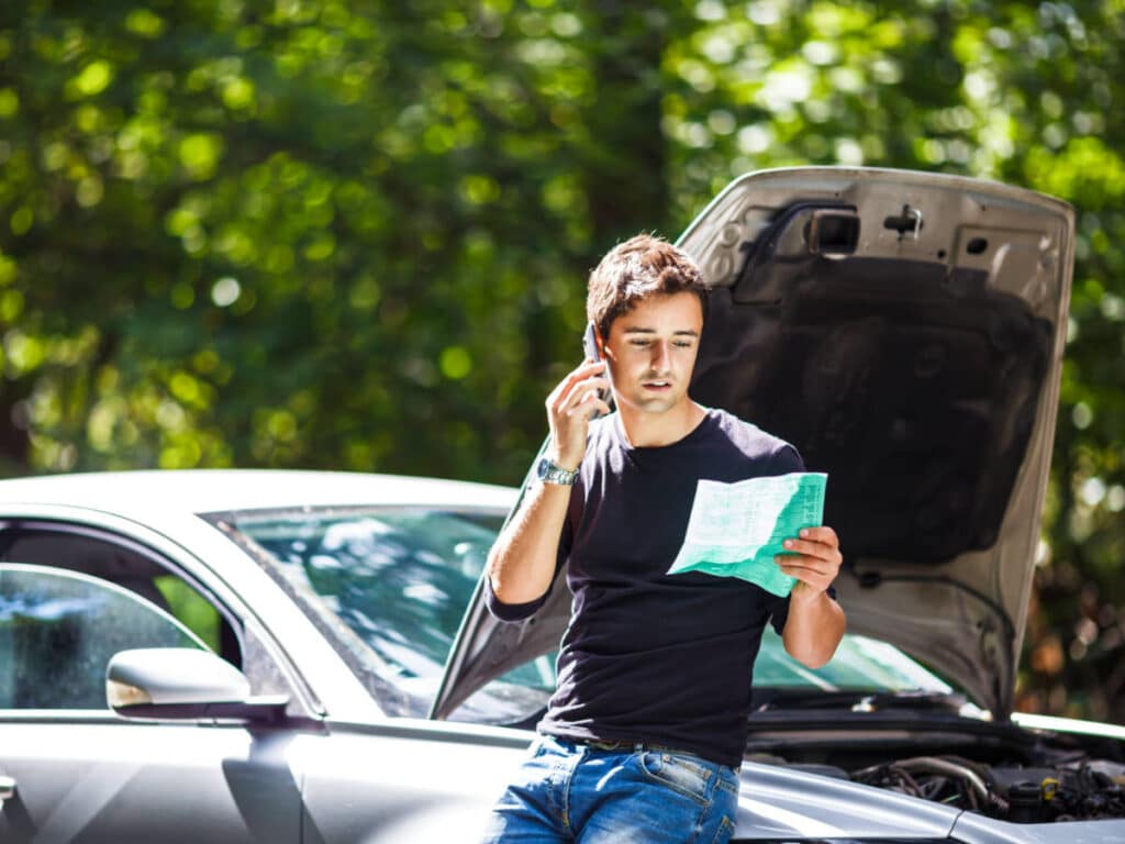 A man sitting on his broken car while calling on his phone and holding a paper