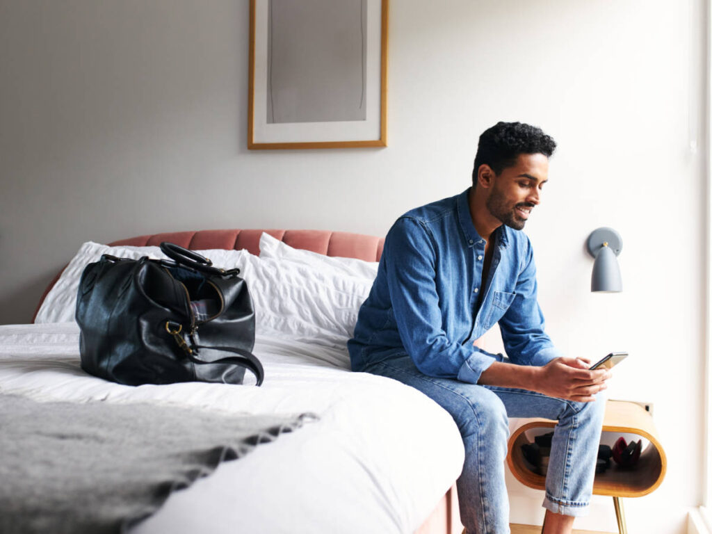 A man looking at his phone while on her bed on a hotel room