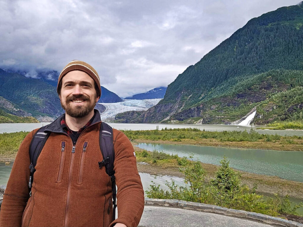 The author Matthew Reppond smiling for a photo with the Mendenhall Glacier and Nugget Falls in the background