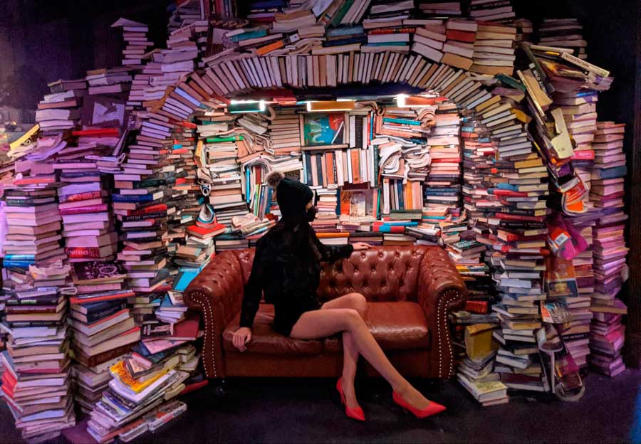 Travel Lemming writer Clara, sitting on a sofa with books on her background in Meow Wolf Convergence Station